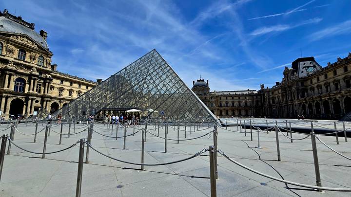 Wide shot of the Louvre Pyramid and surrounding palace buildings under a vivid blue sky.