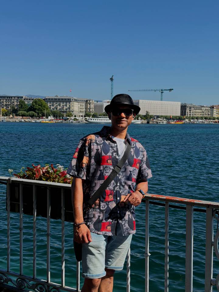 A man wearing sunglasses and a hat poses by the lakeside promenade with Geneva’s urban skyline and blue water behind him.