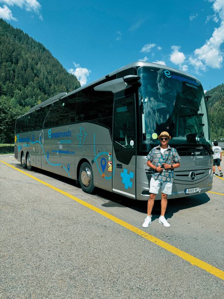 Man posing beside a modern tour coach in a parking area surrounded by forested hills.