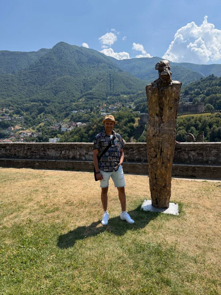 Traveller standing on a grassy terrace with a wooded valley and hilltop fortress ruins in the background.