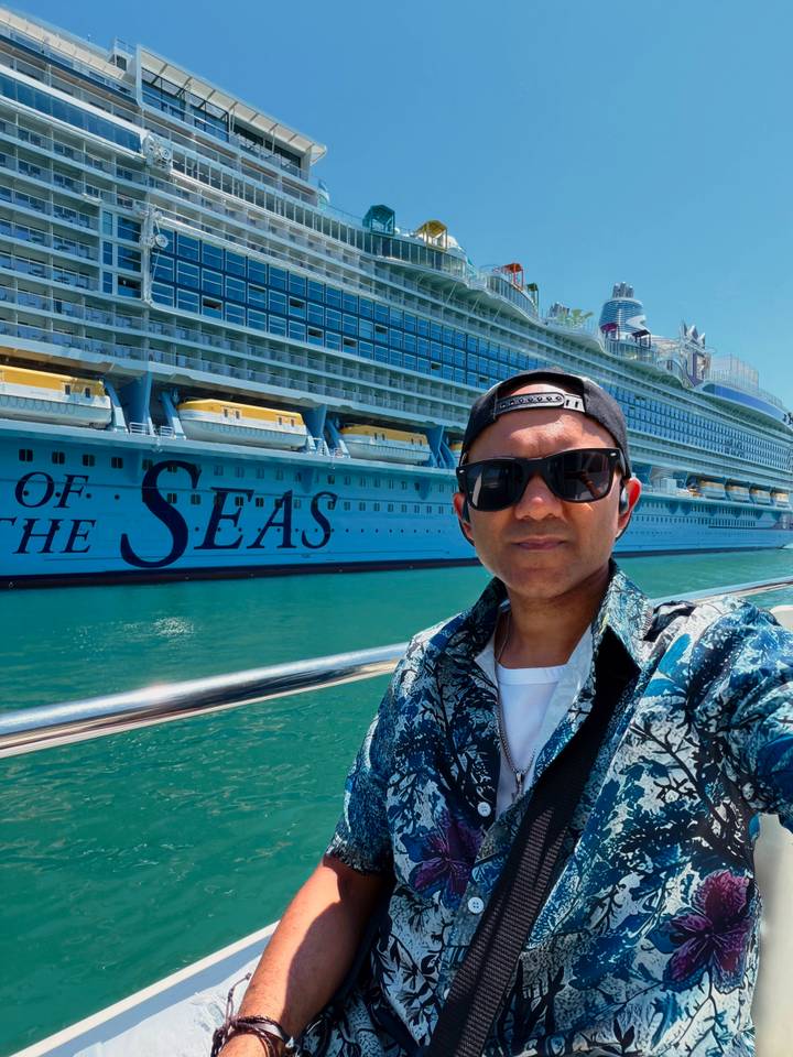 Selfie with a large cruise ship bearing the words “OF THE SEAS” anchored in bright turquoise water.