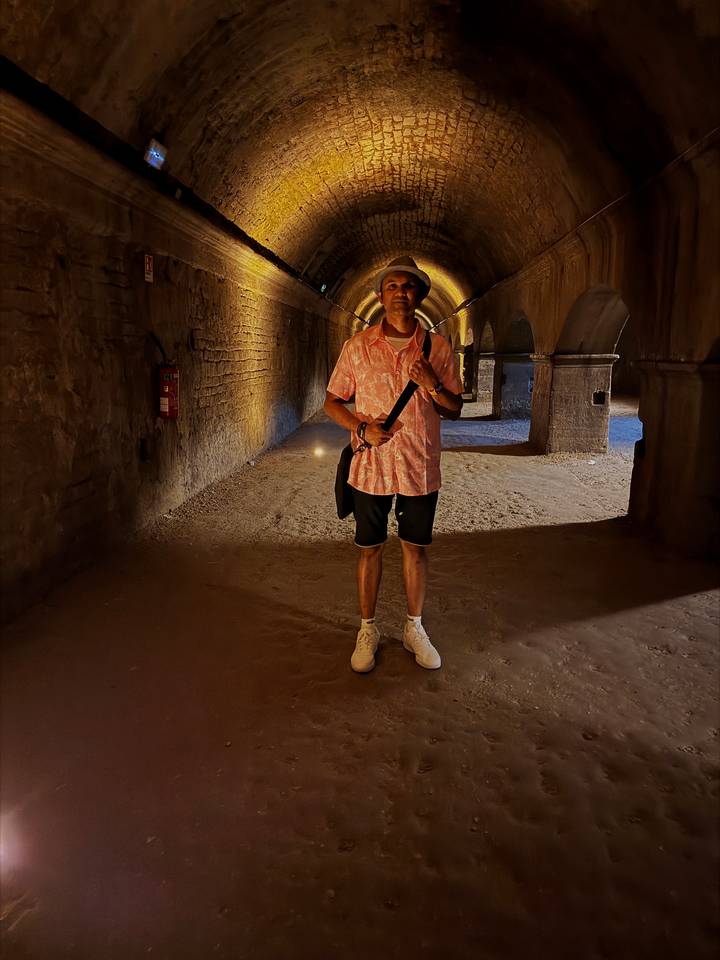 A dim, warm-lit corridor view with a lone traveller standing between stone arches underground.
