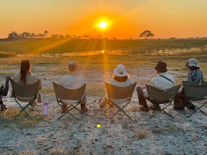 Travellers seated in camp chairs watch the orange sun set over the wetlands, rays streaming across.