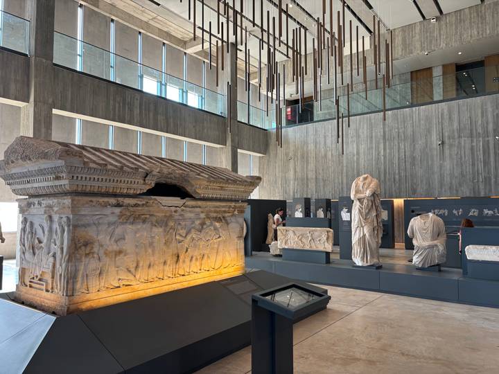 Modern museum hall displaying an ornate sarcophagus and classical marble statues under skylights.