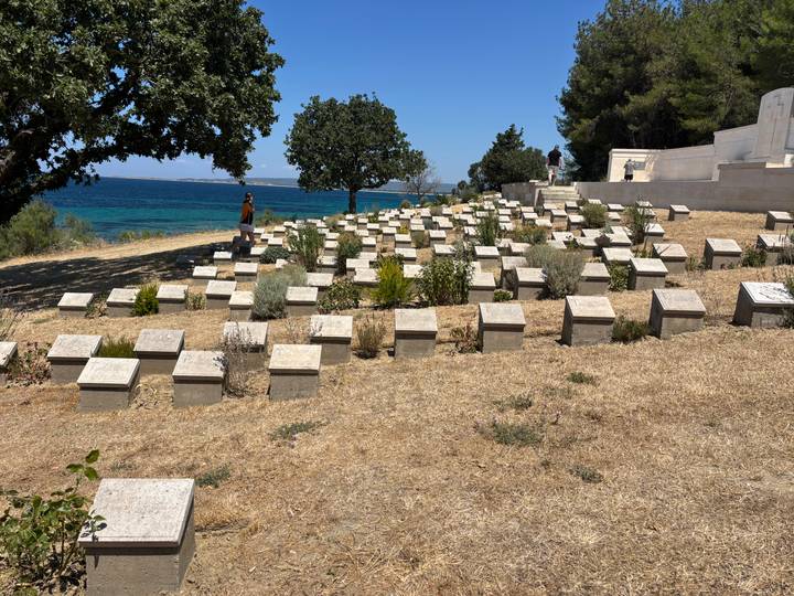 Rows of low stone grave markers overlook a bright blue sea at a coastal war cemetery.