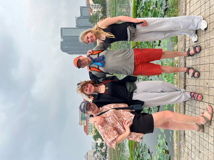 Four travellers pose arm-in-arm beside a lotus pond with modern city high-rises in the background.