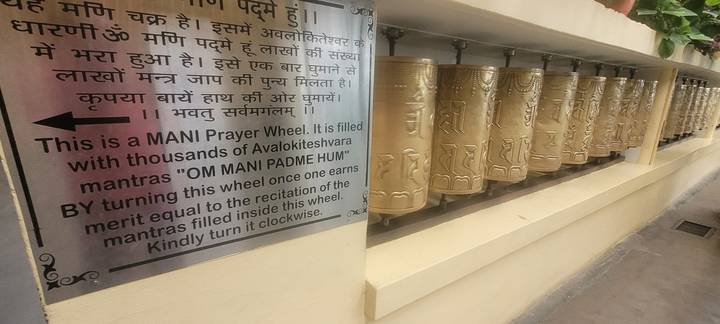 Row of brass Tibetan prayer wheels with bilingual explanatory sign nearby.