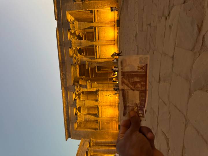Blurry foreground hand holding a 50-pound note against a lit temple facade at dusk.