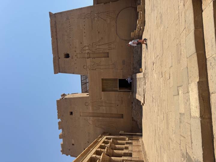 Massive sandstone pylon entrance of Edfu Temple with faint carved figures under bright sky.