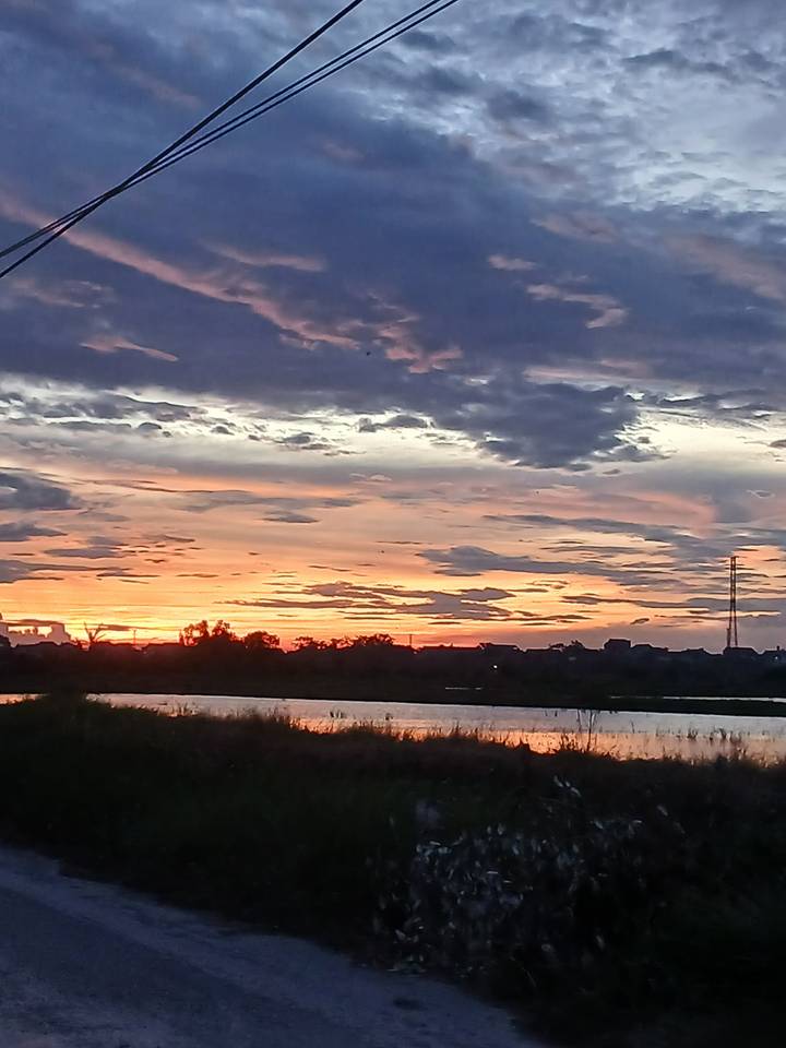 Colourful sunset with dramatic layered clouds reflected on a calm river beyond a distant village silhouette.