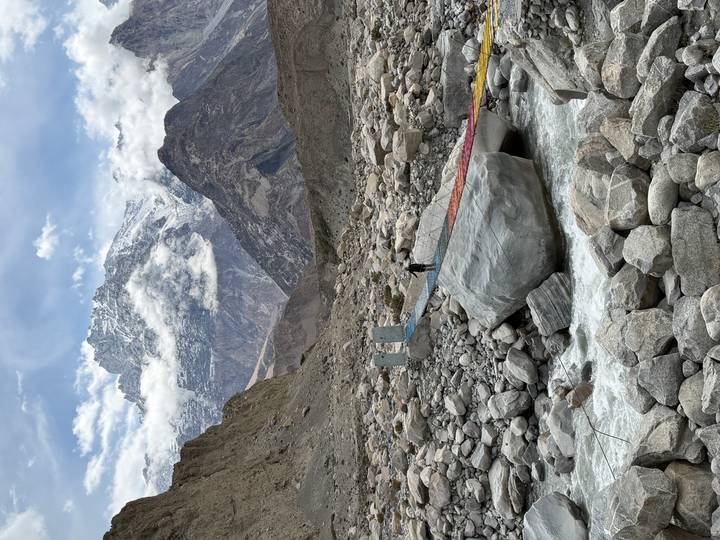 Rainbow-planked suspension bridge crossing a rocky glacial stream with soaring snowy peaks beyond