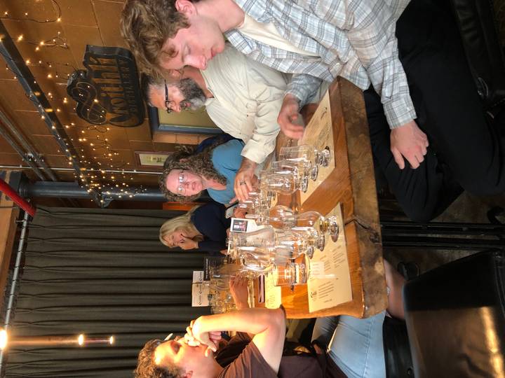 Travelers sit at a rustic distillery table sampling multiple whiskey glasses under warm indoor lights.