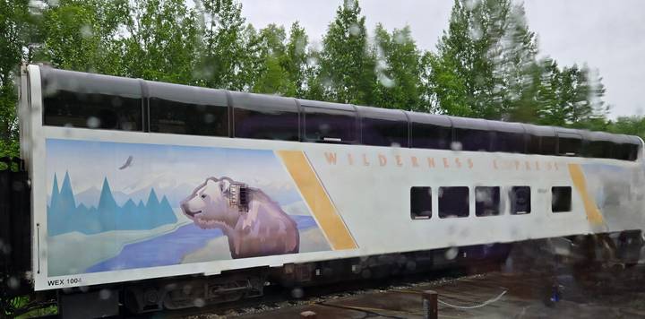 Double-deck Wilderness Express train car viewed through a rain-streaked window, featuring a painted bear and mountain scene.