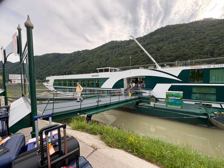 Passenger boarding a green-and-white river cruise ship moored beside wooded hills.