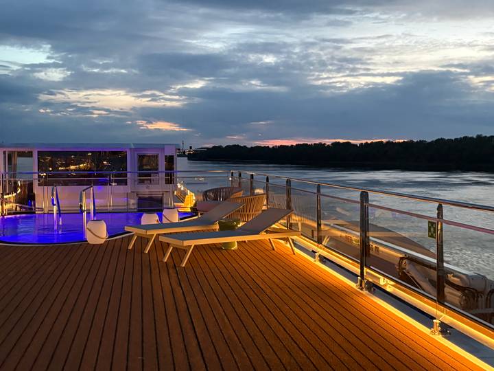 Sunset view from a cruise-ship sundeck with loungers, pool lights and a calm river.
