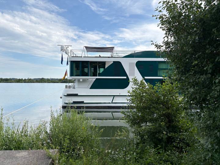 Modern river ship docked beside calm water and green shoreline under a blue sky.