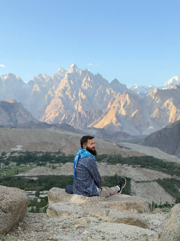 Bearded traveler wrapped in a scarf with the jagged Passu peaks towering behind him.