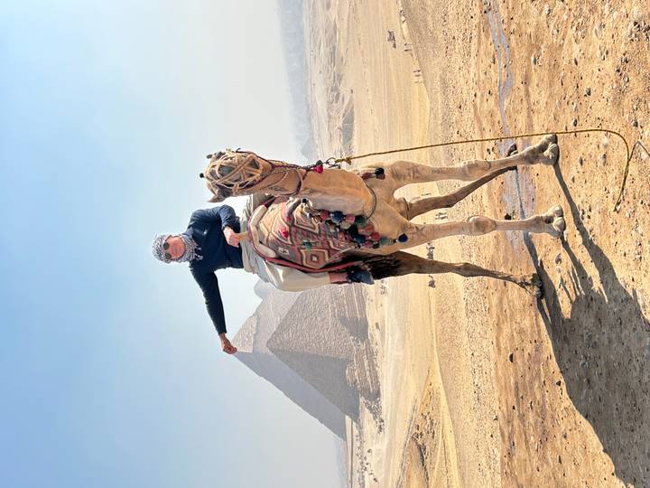 Traveller atop a decorated camel strikes a pose with the Great Pyramid rising through desert haze behind.