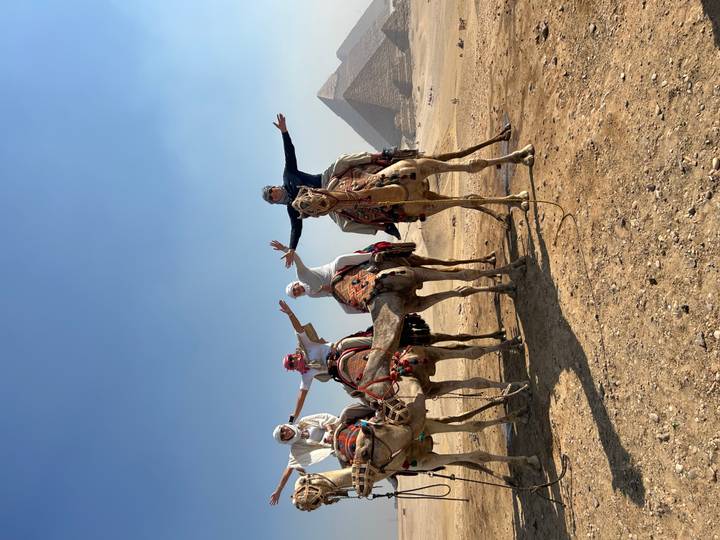 Group of riders atop camels raise their arms in triumph with the pyramids looming in the background.