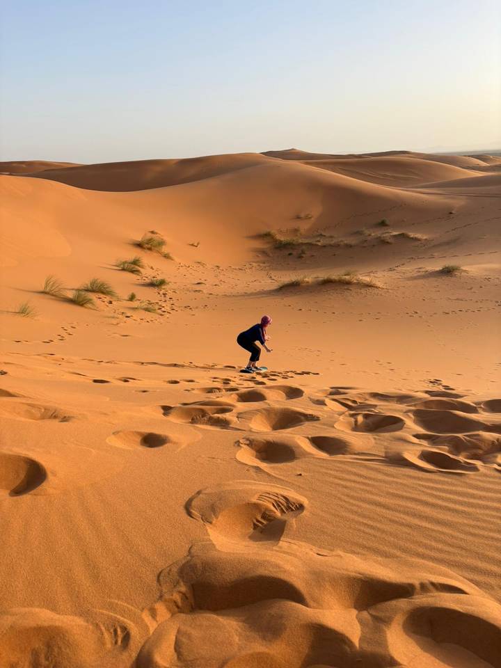 Woman clambers up an orange dune in the Sahara, footprints trailing behind in rippled sand.