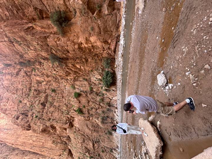 Travellers photograph towering red rock walls beside a shallow stream in Todra Gorge.