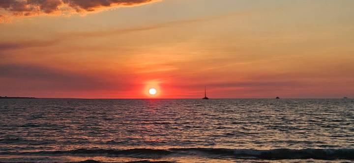 Brilliant orange sun setting over rippling ocean water with a small sailboat silhouetted on the horizon