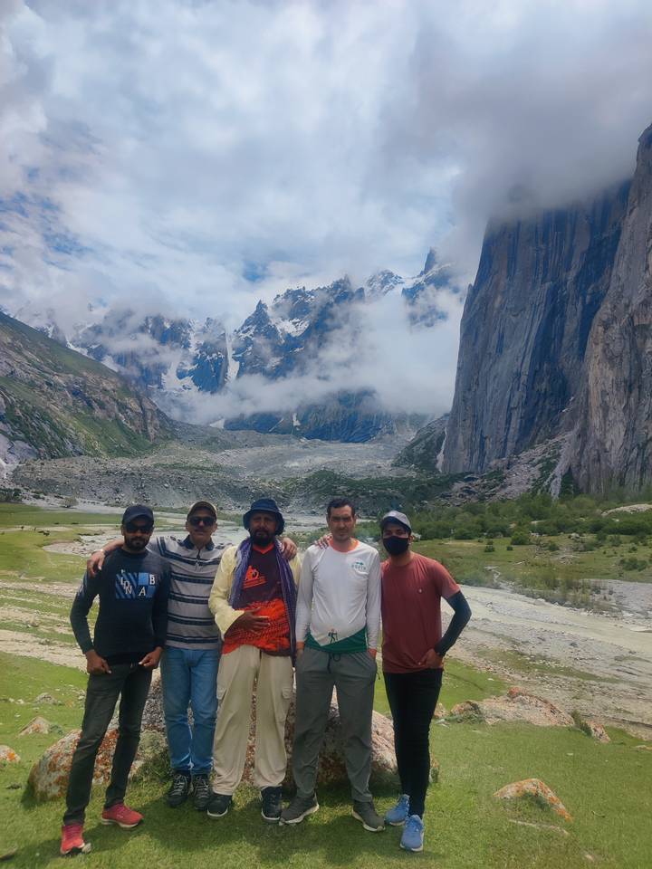Five trekkers posing together in a dramatic alpine valley backed by jagged snow-capped peaks and misty clouds.