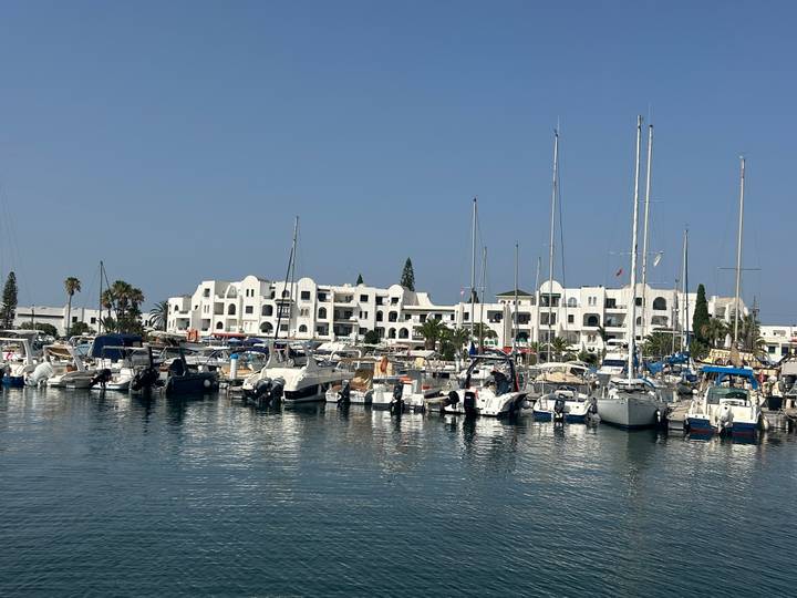 Marina filled with sailboats and motor yachts in front of white Moorish-style resort buildings under a clear sky.