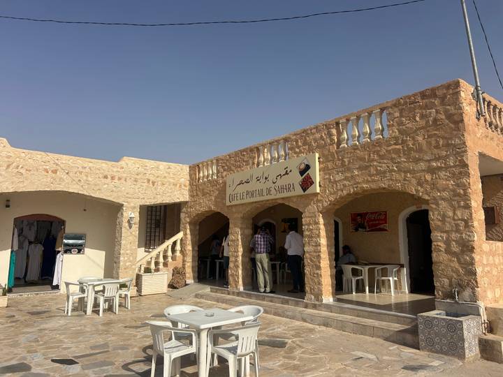 Stone-built roadside cafe with shaded terrace and Arabic signage offering refreshments in the Tunisian desert heat.