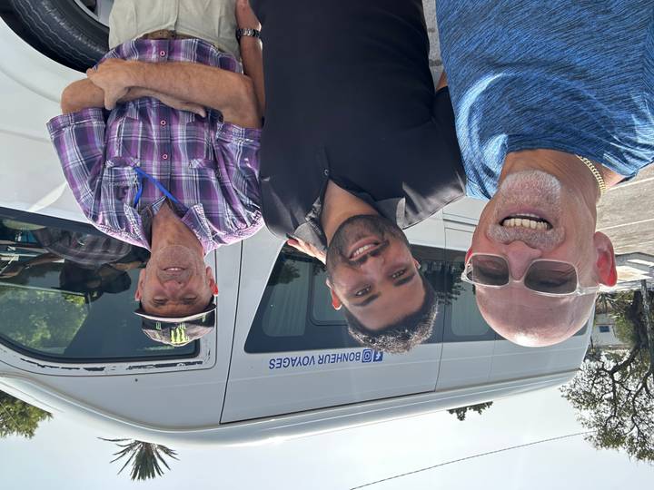 Three smiling men pose for a selfie in front of a white tour van branded Bonheur Voyages.