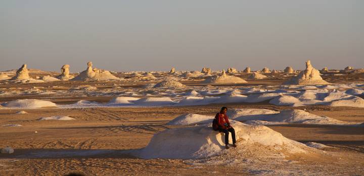 Lone traveler seated on a chalky mound amid surreal white rock formations stretching to the horizon.
