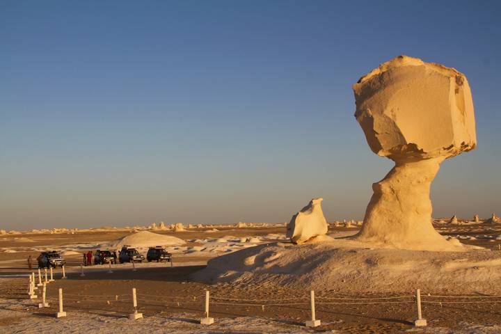 Massive chalk monolith dominating a desert scene with distant 4x4 vehicles and golden evening light.