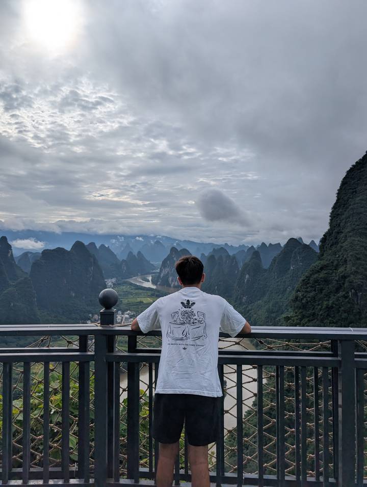 Traveller gazes out over dramatic karst mountains and winding river valleys under a cloudy sky.