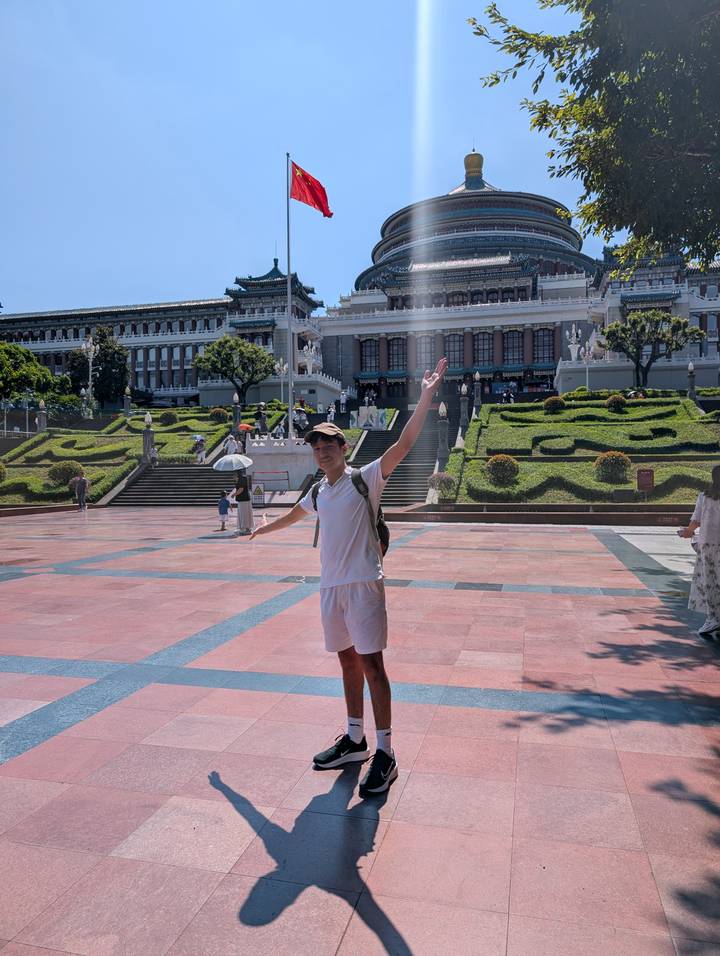 Happy traveller spreads arms in front of grand neoclassical hall and manicured gardens on a sunny day.