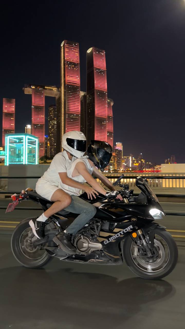 Two riders in white helmets speed across a lit city bridge on a motorbike at night.