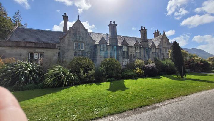 Historic manor house with tall chimneys and stone façade surrounded by manicured lawn and shrubs on a bright day.