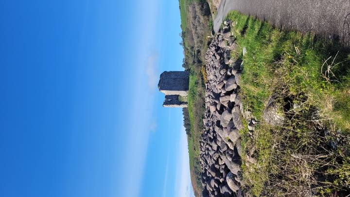 Ruined stone tower standing on a grassy rise with boulder field and winding path under a vivid blue sky.