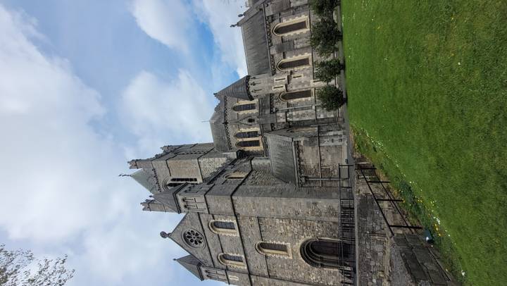Impressive grey-stone Christ Church Cathedral towering above a green lawn under broken clouds.