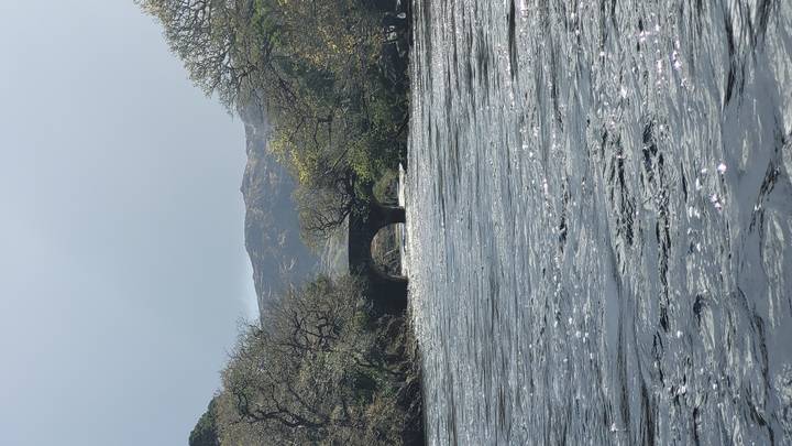 Wide river with fast-flowing water and an old stone arch bridge framed by spring trees and distant hills.