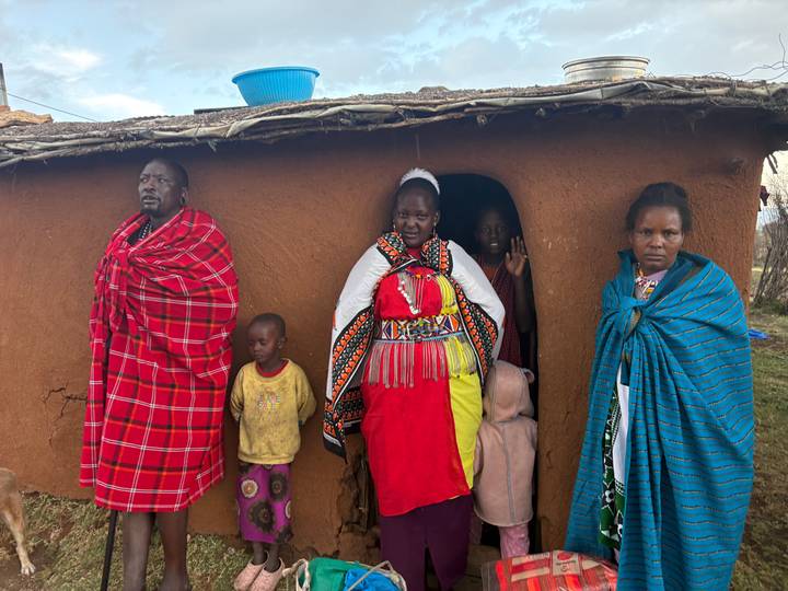 Maasai family dressed in traditional shukas and beadwork standing at entrance of mud hut.