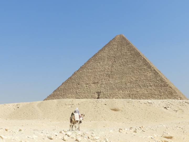 Desert scene with lone camel rider approaching a massive pyramid under clear sky