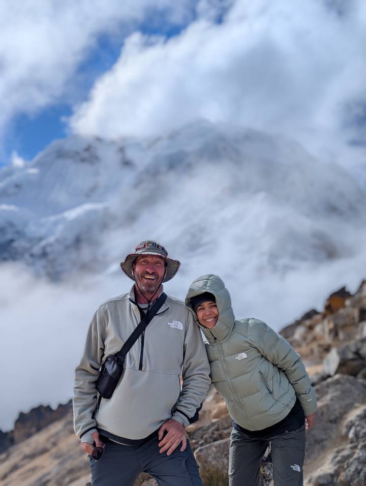 Smiling couple bundled in jackets stands before a misty snow-covered Andean peak.