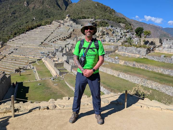 Hiker in neon green shirt stands proudly on Machu Picchu terraces with stone ruins cascading behind.