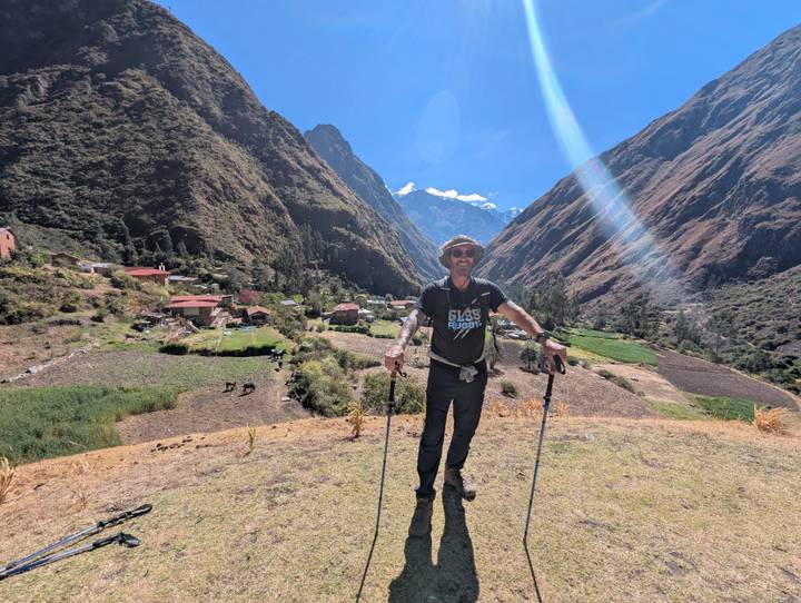 Trekker with poles spreads arms in a lush Andean valley village with sun flare across the lens.