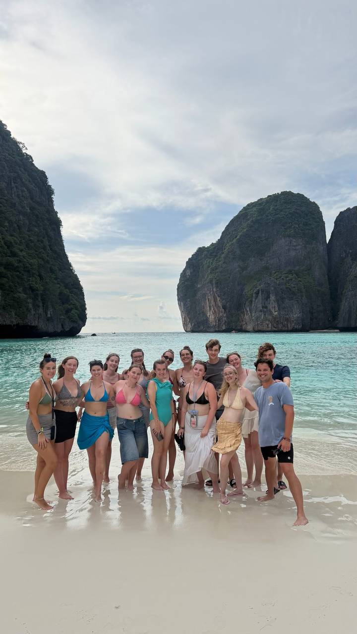 Cheerful tour group in swimwear posing on a turquoise bay framed by dramatic limestone cliffs and white sand.