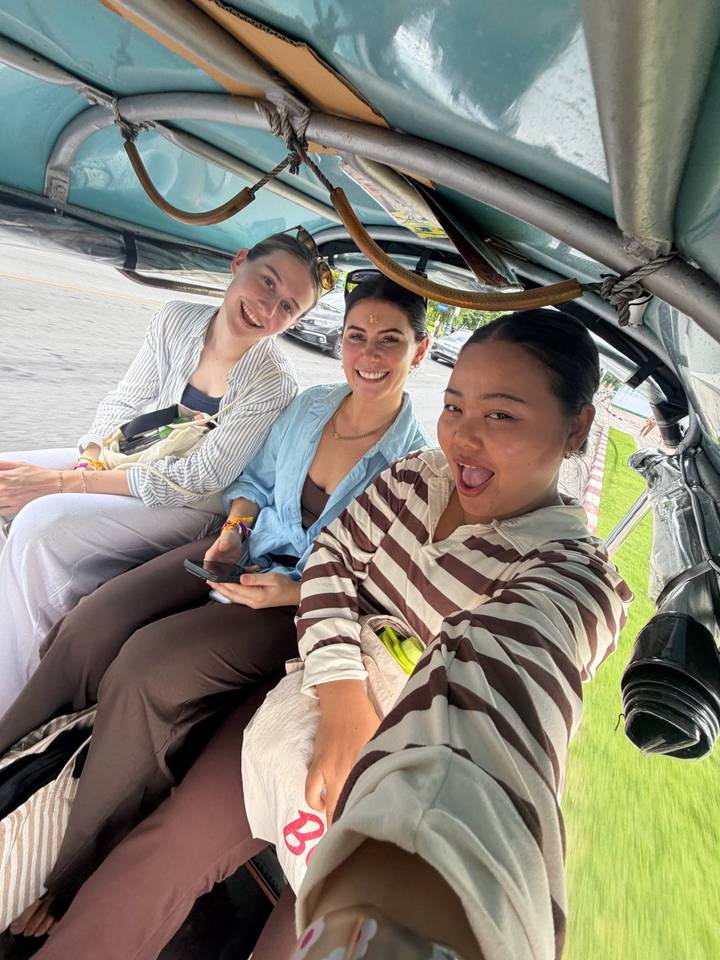 Three friends smiling for a selfie while riding in an open tuk-tuk on a city street.