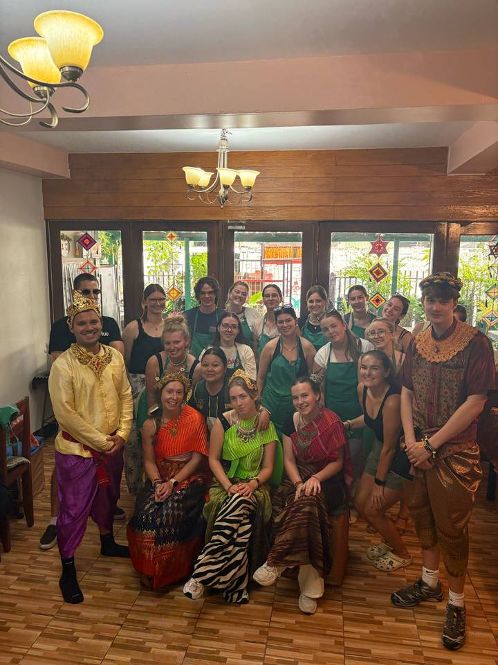 Large tour group wearing aprons and Thai cultural outfits posing inside a cooking classroom.