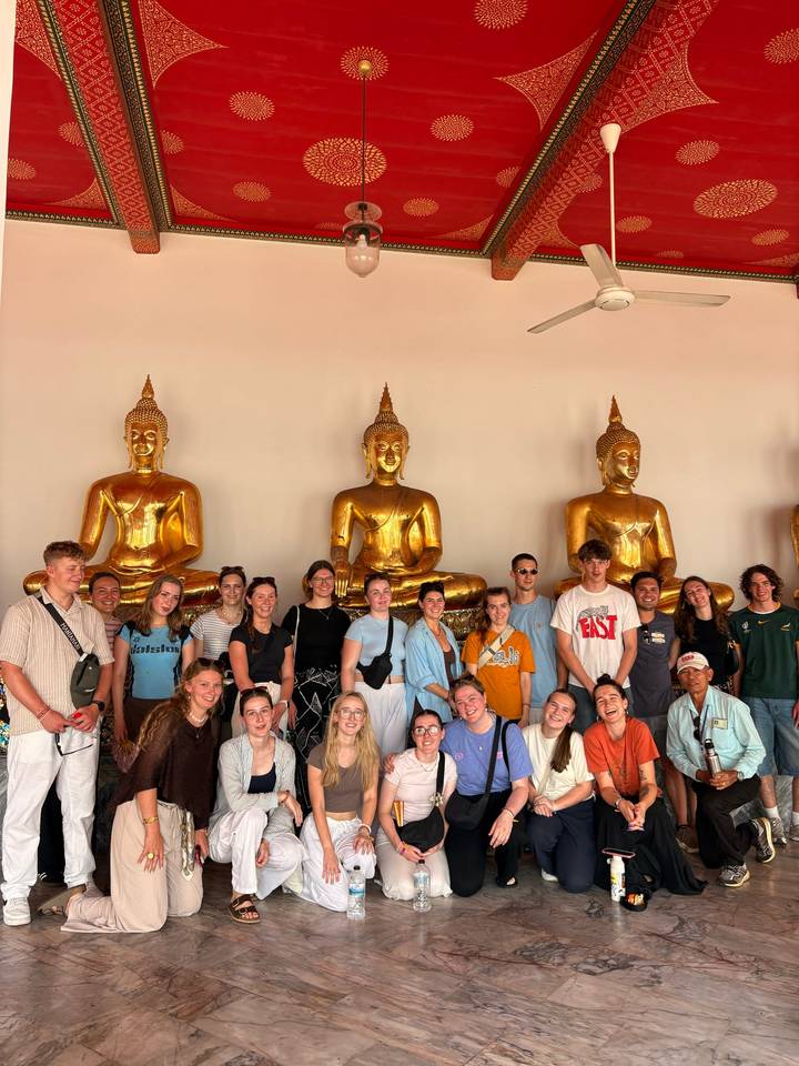 Travel group standing in front of a row of golden seated Buddha statues inside a Thai temple gallery.