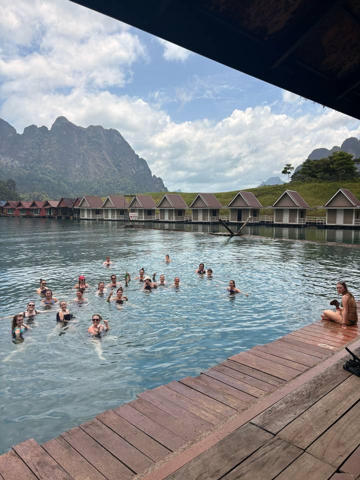 Dozens of travellers swimming in a calm lake with floating bungalow cabins backed by misty limestone mountains.