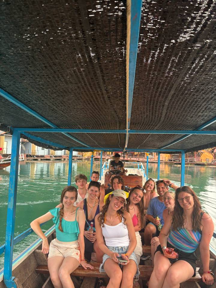 Smiling travellers seated under a canopy on a long-tail boat gliding across emerald water toward floating houses.
